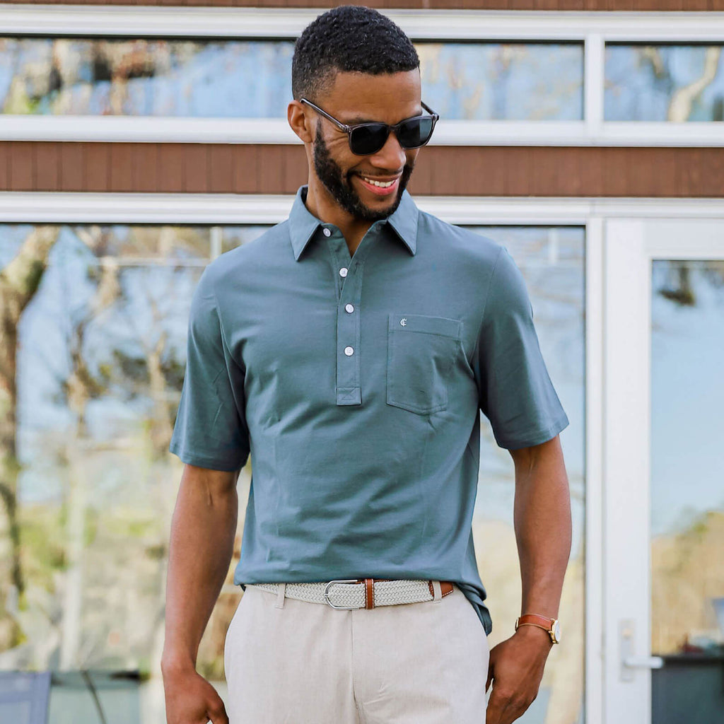 A male model standing outside wearing sunglasses and a light blue polo shirt